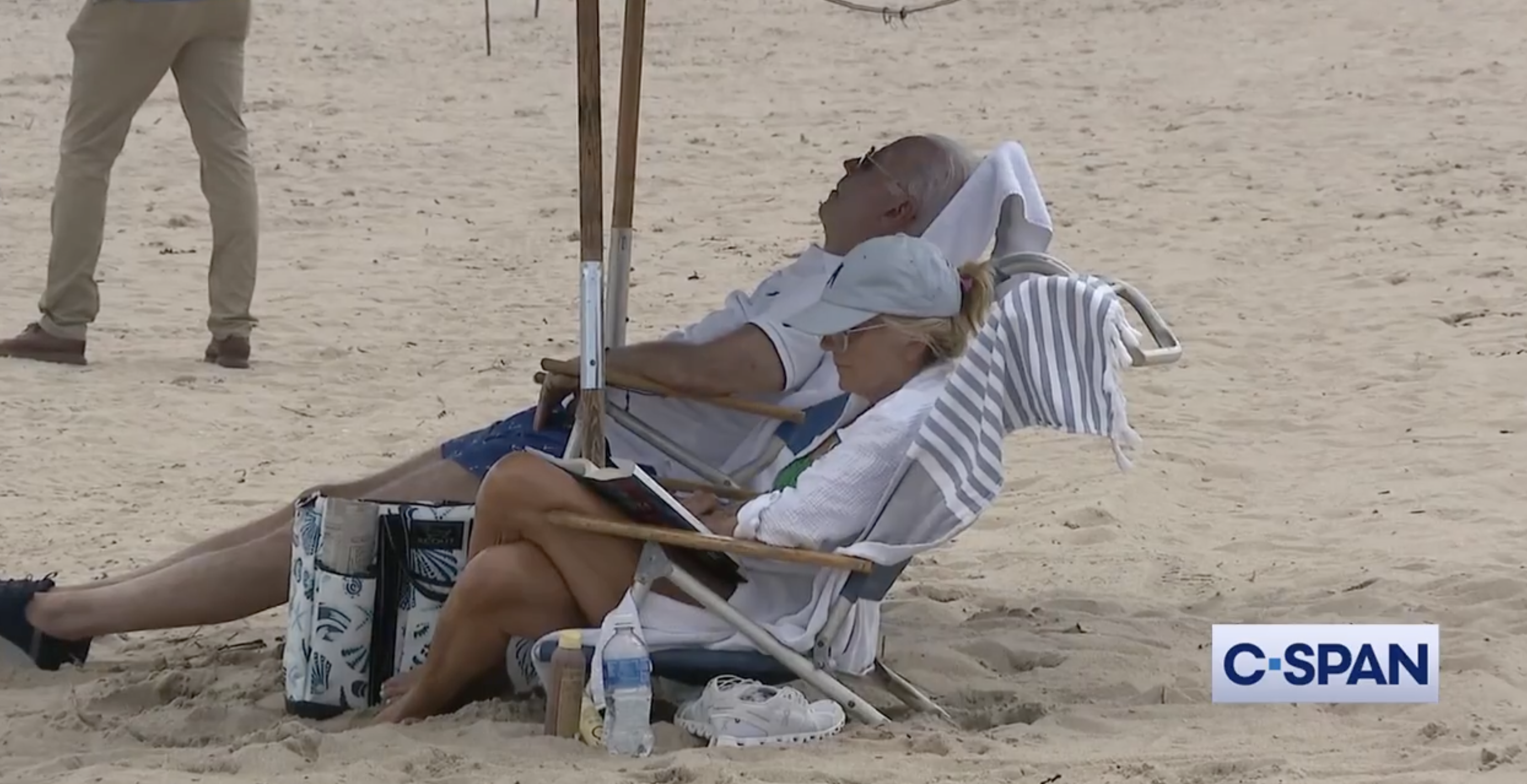 Joe Biden and Jill Biden relax on beach chairs under an umbrella. Joe rests with eyes closed while Jill reads a folder