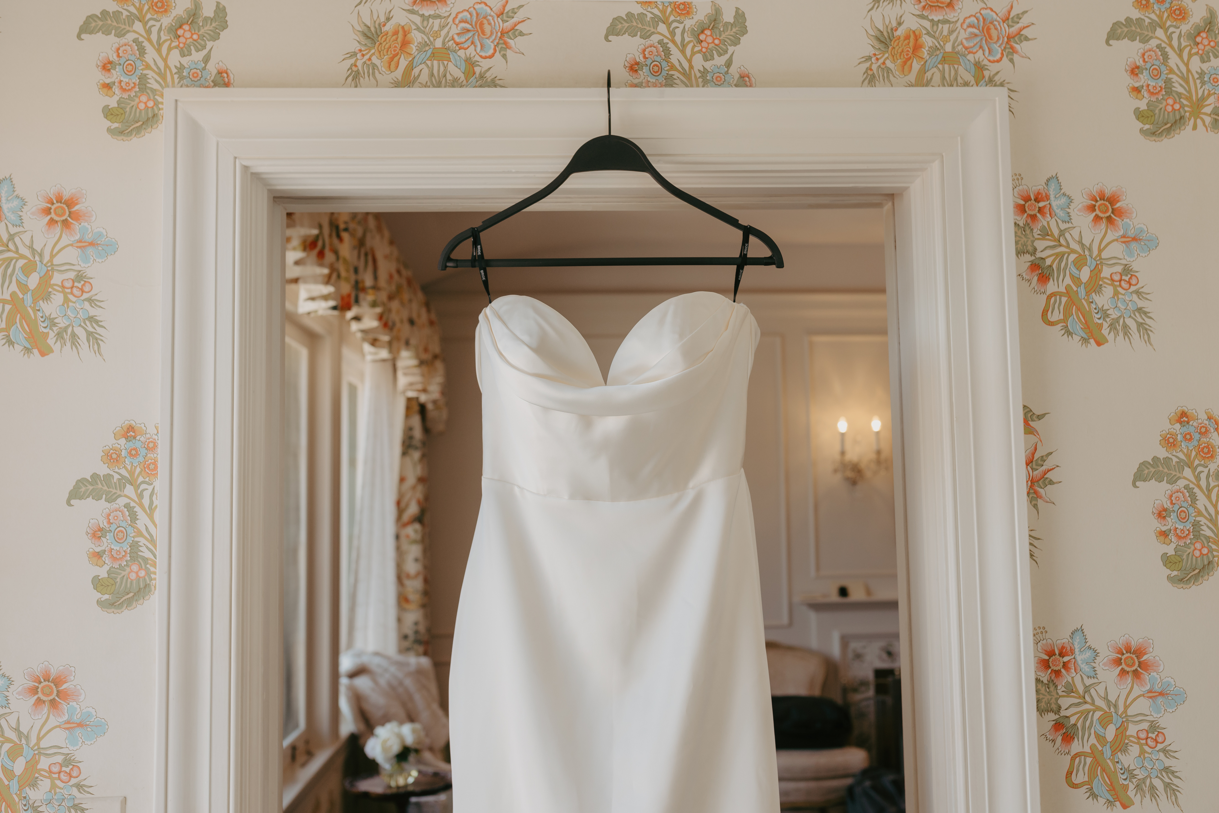 Strapless white wedding dress on a black hanger, displayed against a floral wallpaper background in a room with elegant furnishings