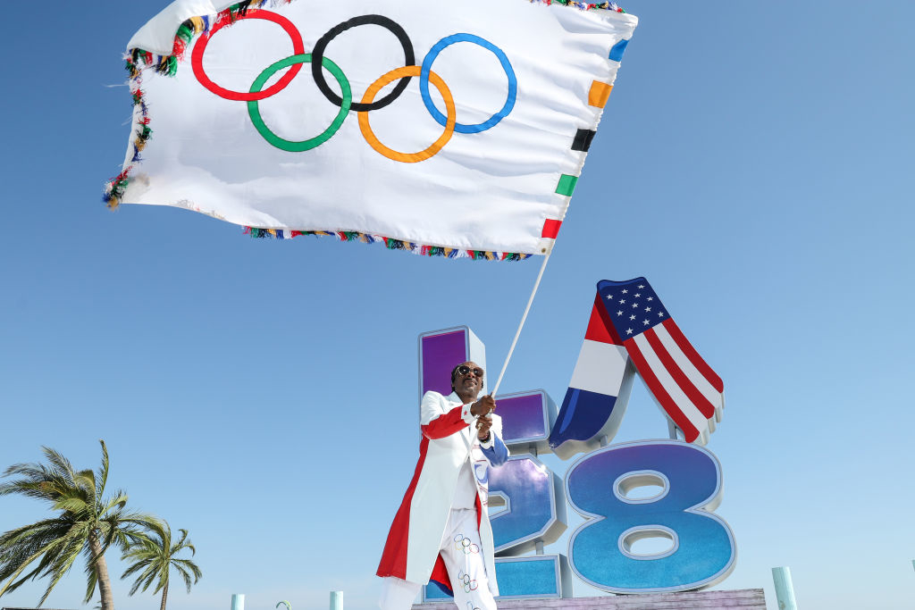Snoop Dogg waves a flag with the Olympic rings in front of a large "LA 28" sign and an American flag