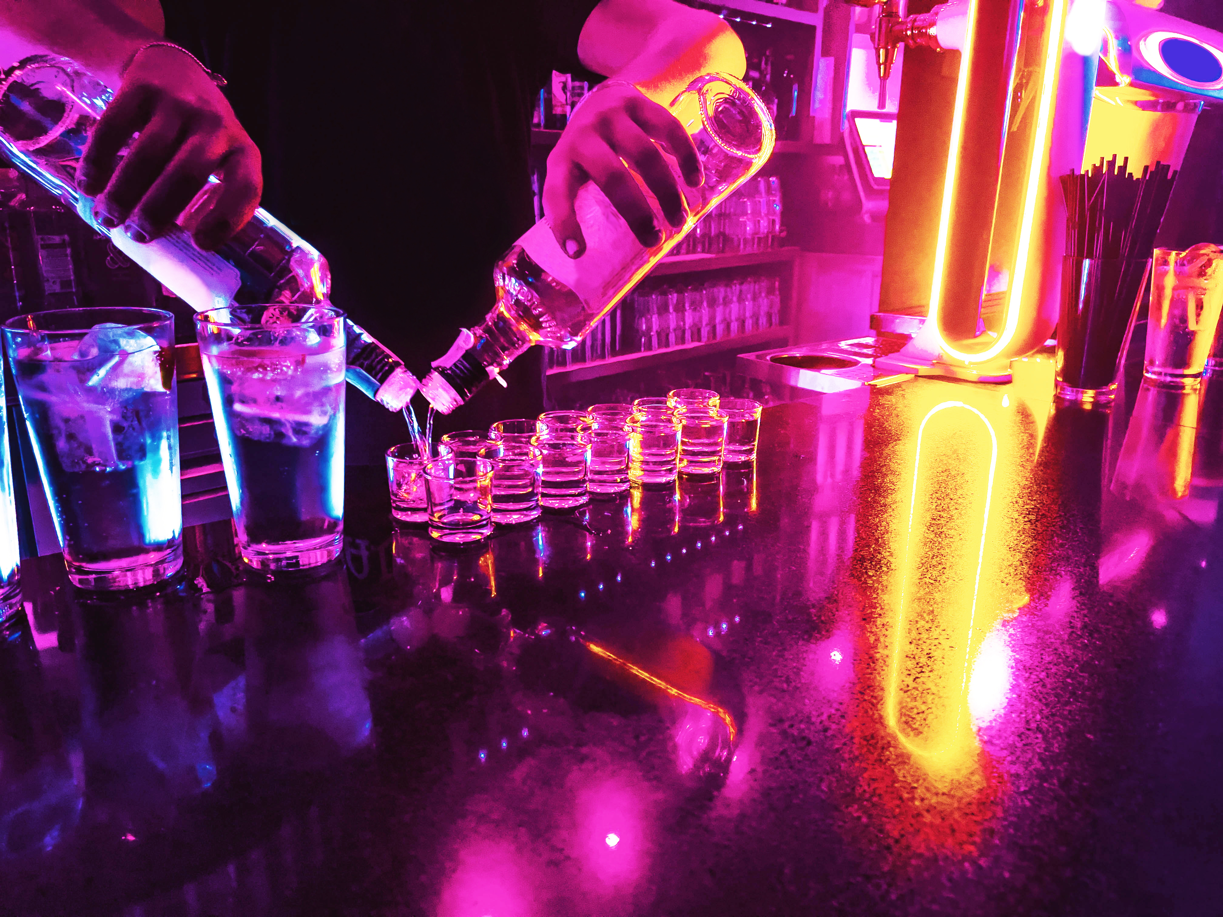 A bartender pours liquor into multiple shot glasses on a neon-lit bar counter