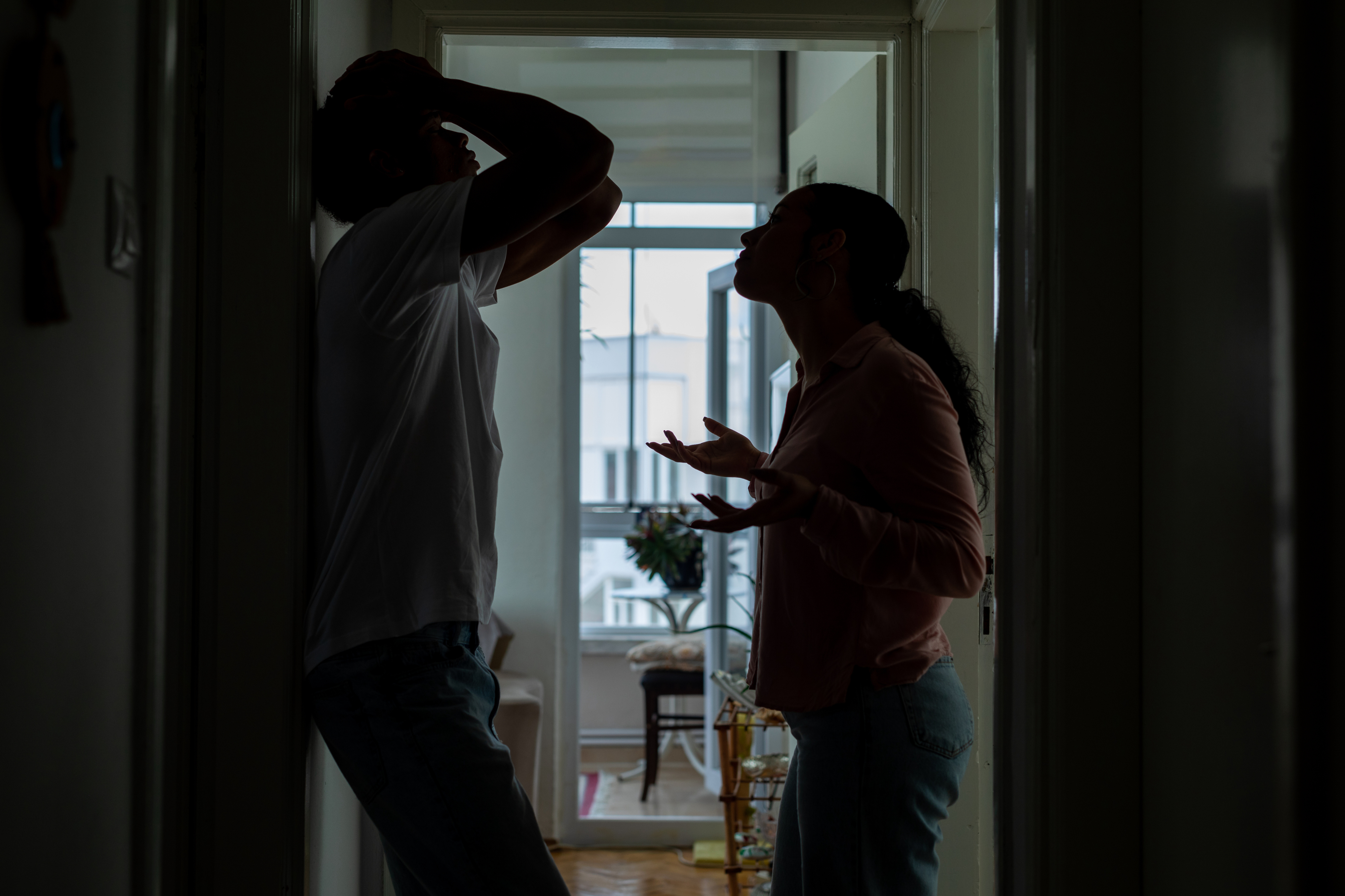 A silhouetted couple argues in a dimly lit hallway; the woman gestures with frustration while the man leans back against the wall holding his head