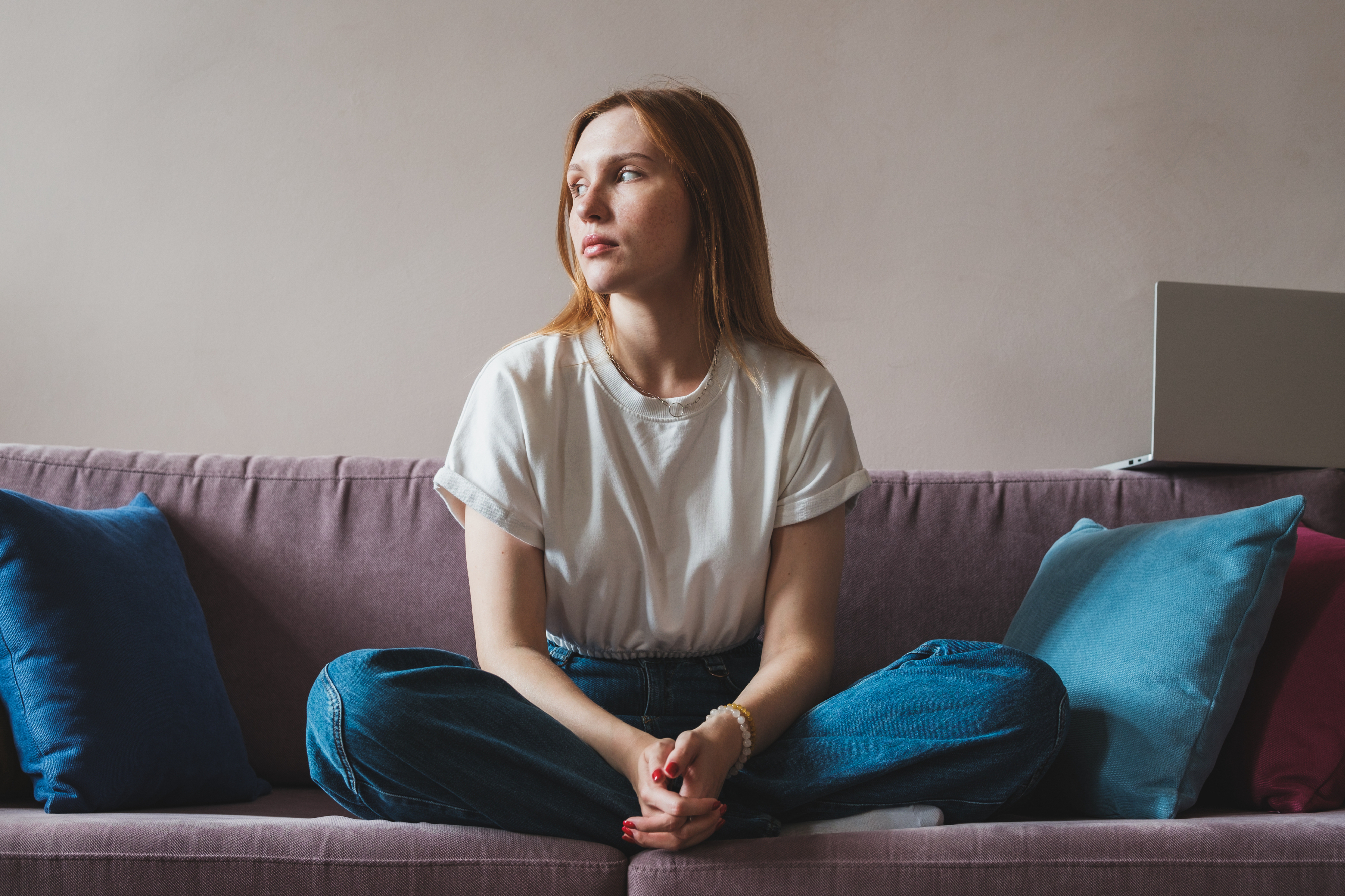 Woman in casual clothing, sitting cross-legged on a couch with a laptop nearby, looking contemplative. No other persons present