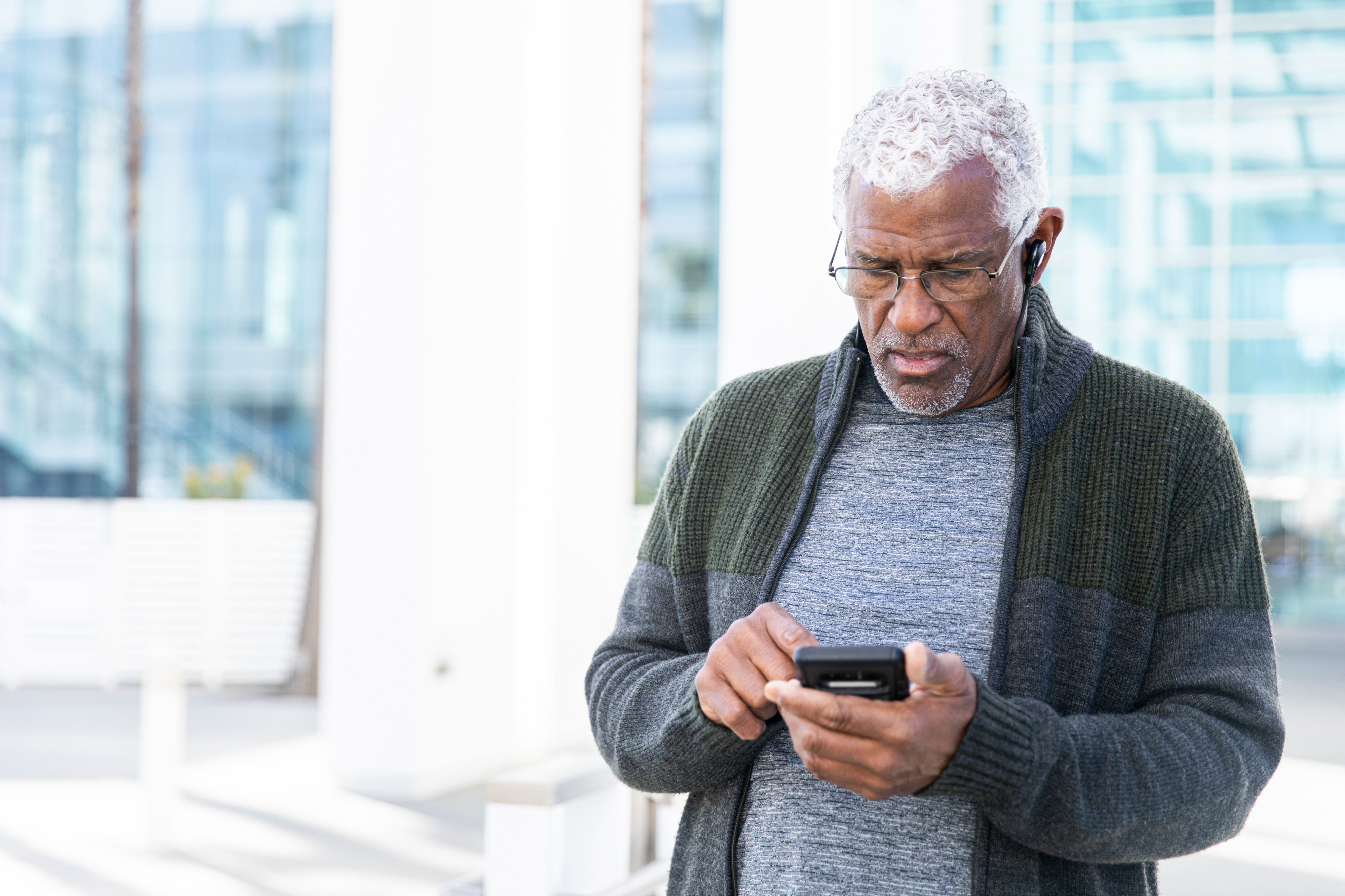 An older man with short curly hair and glasses looks at his phone. He is wearing a sweater and earphones