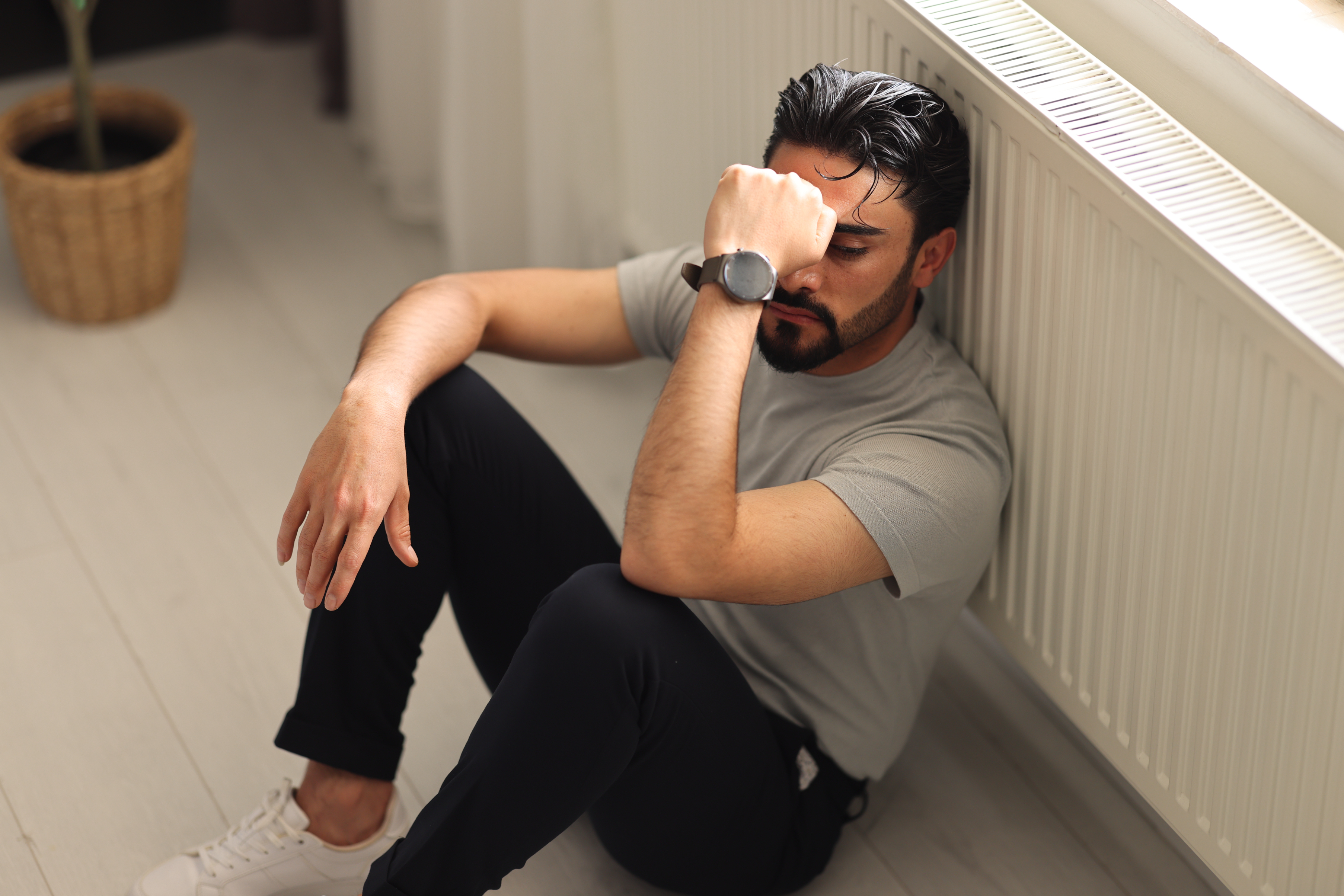 A man in a gray t-shirt and dark pants sits on the floor near a radiator, resting his head on his hand, appearing stressed or tired