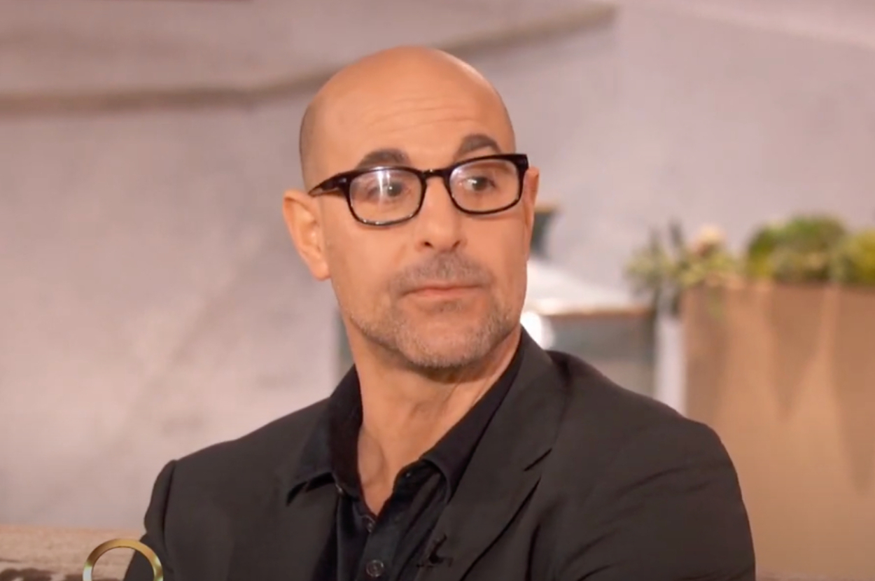 A balding man with glasses, Stanley Tucci, is seen in a still image from an interview. He is wearing a dark blazer and shirt, looking slightly to the side