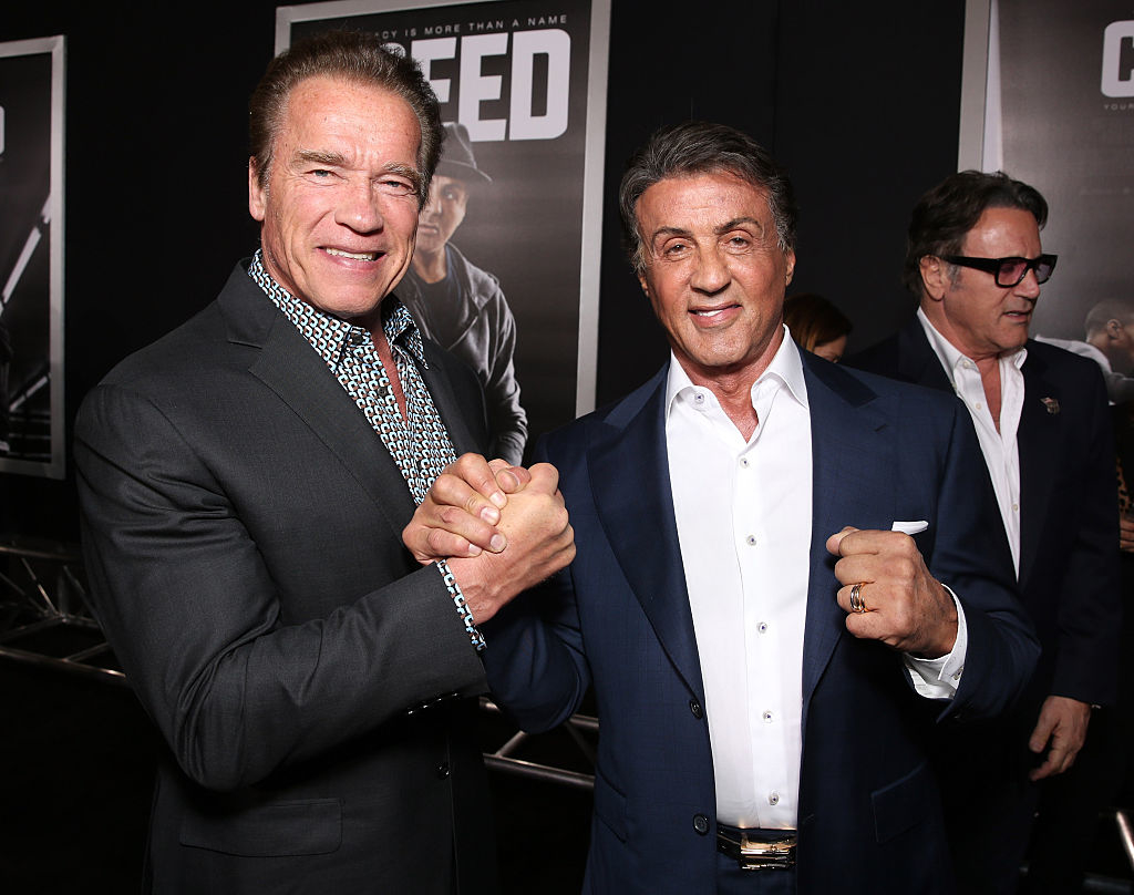 Arnold Schwarzenegger and Sylvester Stallone shaking hands at the premiere of Creed. Both are wearing suits, Arnold in a patterned shirt and Sylvester in a white shirt