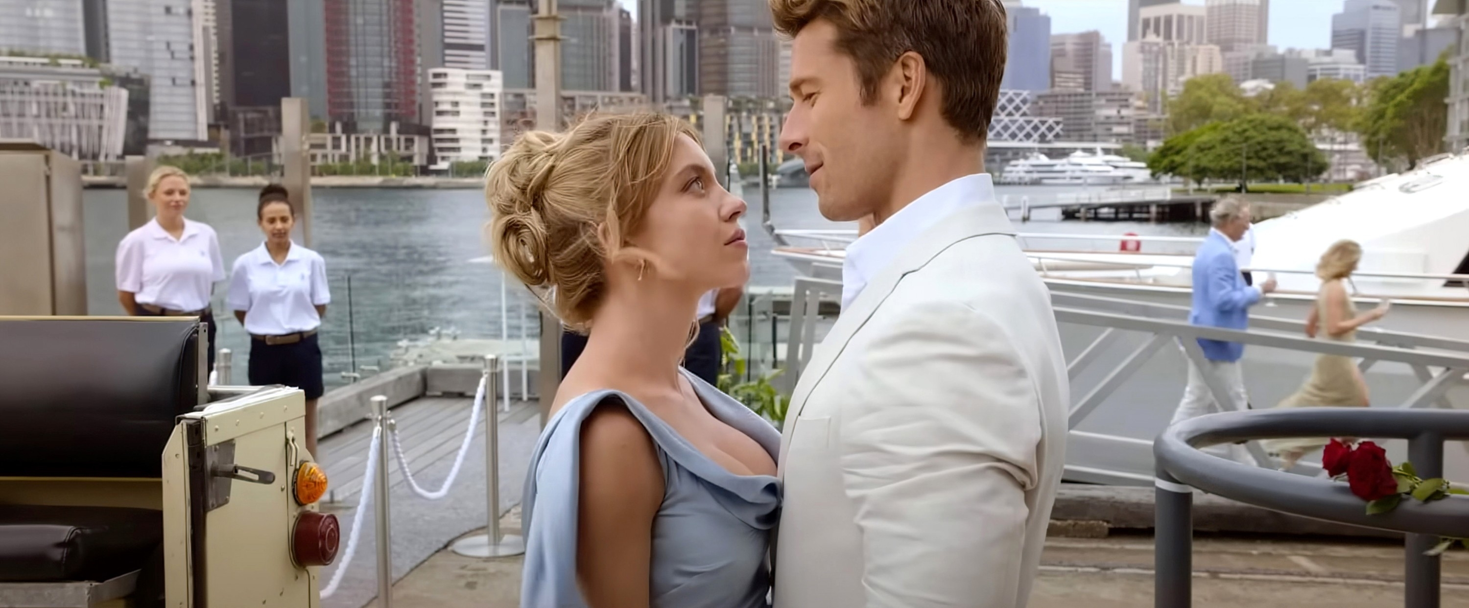 Sydney Sweeney and Glen Powell in character, share an intimate moment on a pier with a cityscape backdrop