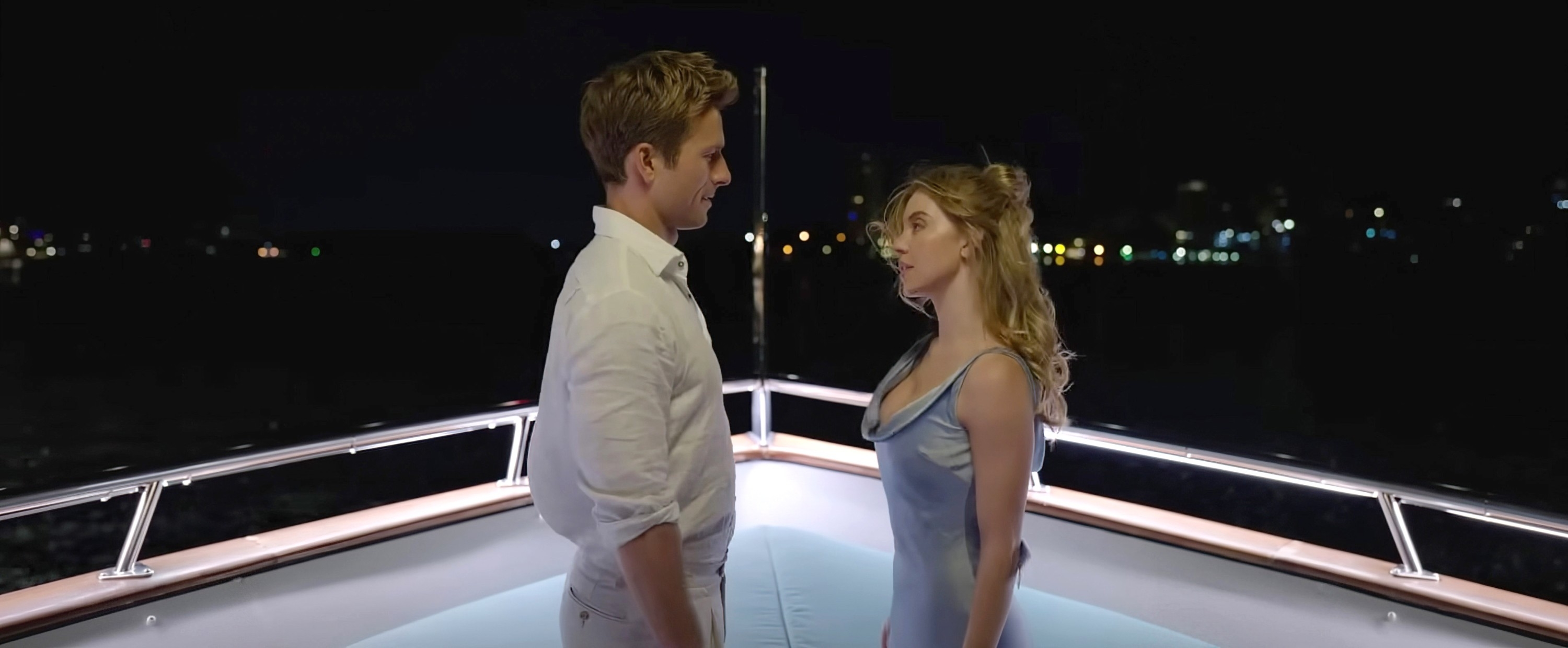 Glen Powell and Syndye Sweeney stand on a yacht at night, facing each other. Lily James is wearing a sleek satin dress, and they appear to be in a serious conversation
