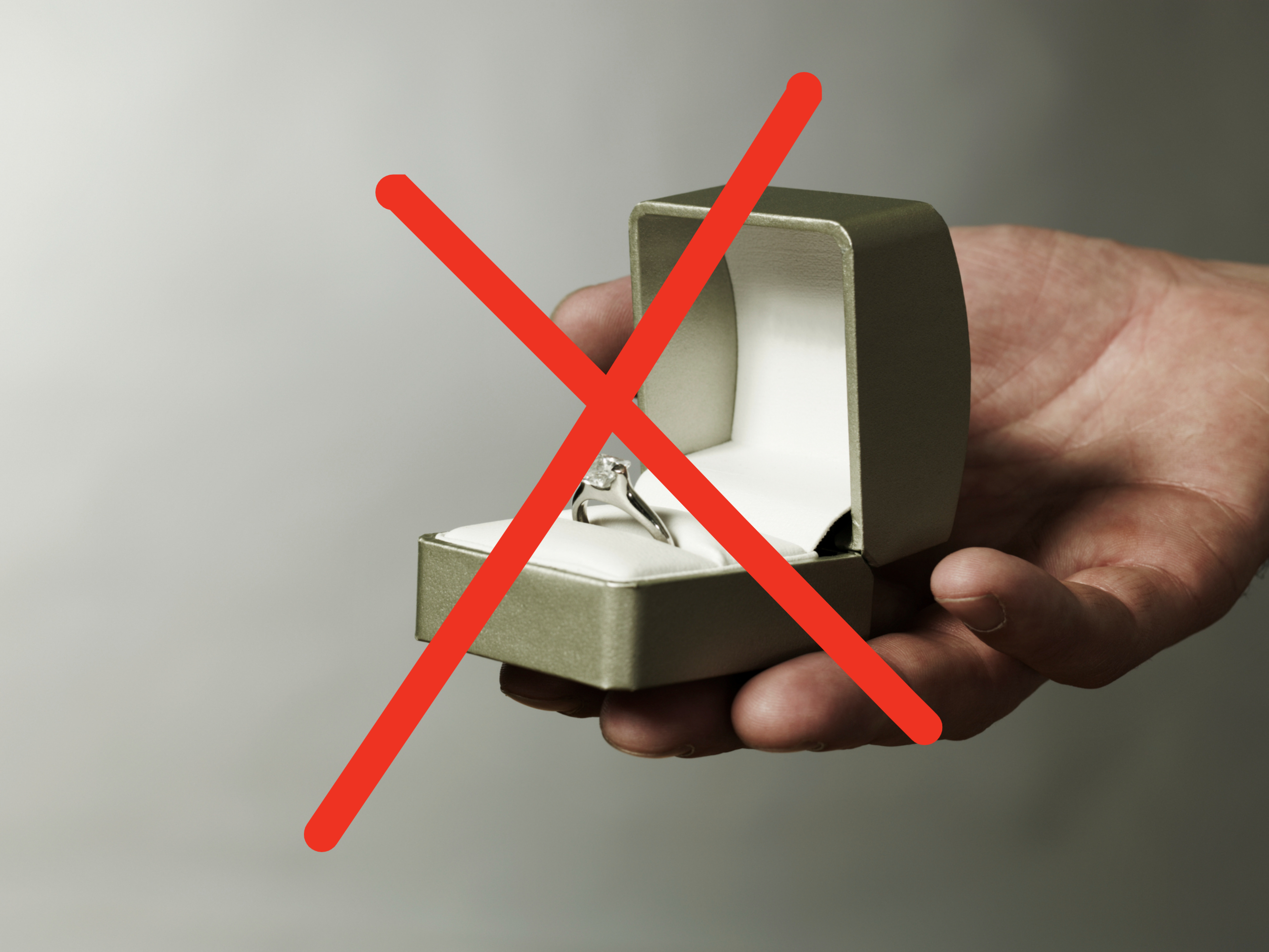 A hand holding an open ring box with an engagement ring inside. The box is greenish-grey and white on the inside