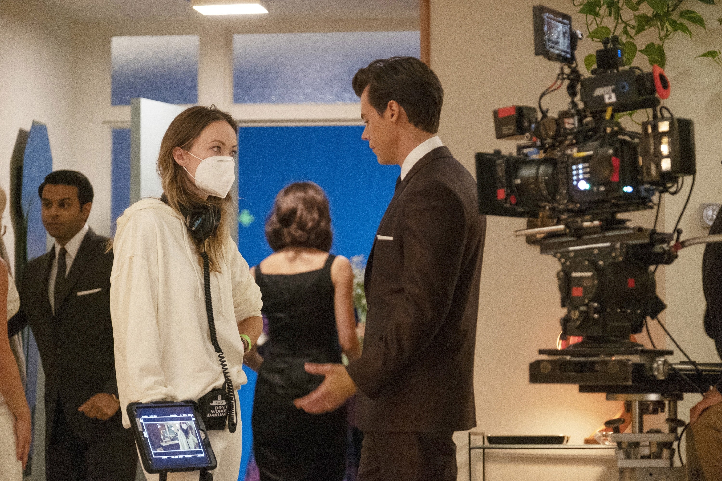 Olivia Wilde, in a casual outfit and mask, directs Harry Styles, in a suit, on a set with cameras and crew members around