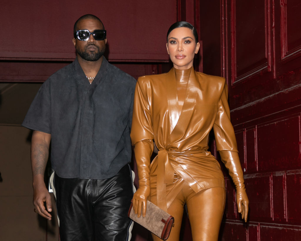 Kanye West in a dark outfit and sunglasses, standing next to Kim Kardashian, who is dressed in a sleek, shiny tan leather ensemble, holding a clutch
