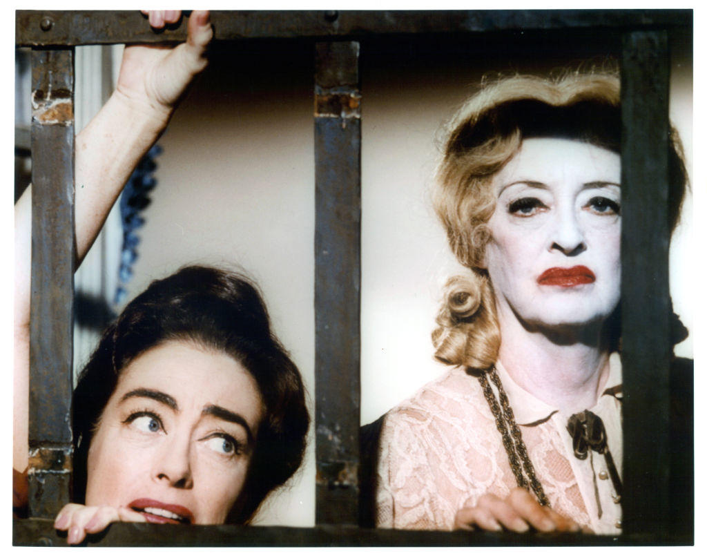 Joan Crawford and Bette Davis look through iron bars; Davis wears a lace top with a bow tie, while Crawford peers with a wary expression