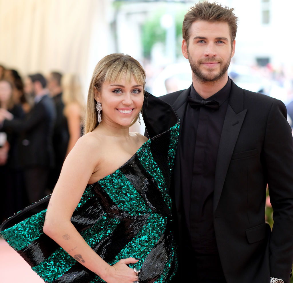 Miley Cyrus in a modern, asymmetrical dress with green sequins stands beside Liam Hemsworth in a black suit and bow tie at a formal event