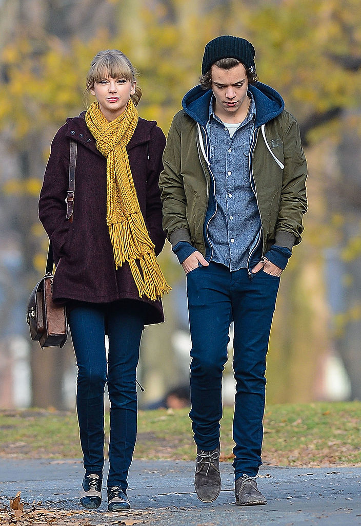 Taylor Swift and Harry Styles walking outside, dressed in casual fall clothing. Taylor wears a scarf and coat, and Harry wears a beanie, jacket, and boots