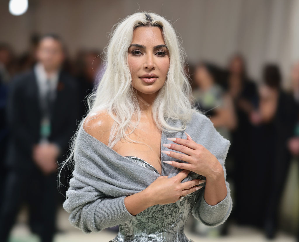 Kim Kardashian at a formal event, wearing a strapless, metallic dress with a shawl draped over her shoulders, posing for the camera. People stand in the background