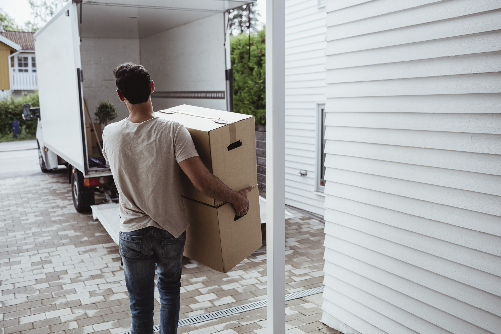 A man carries a large cardboard box from a parked moving truck into a house while wearing a casual T-shirt and jeans