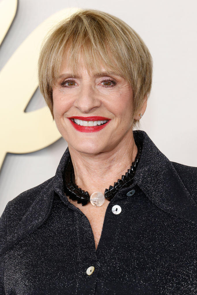 Patti LuPone, smiling with a short hairstyle, wears a shiny, dark top and a black necklace with a clear pendant