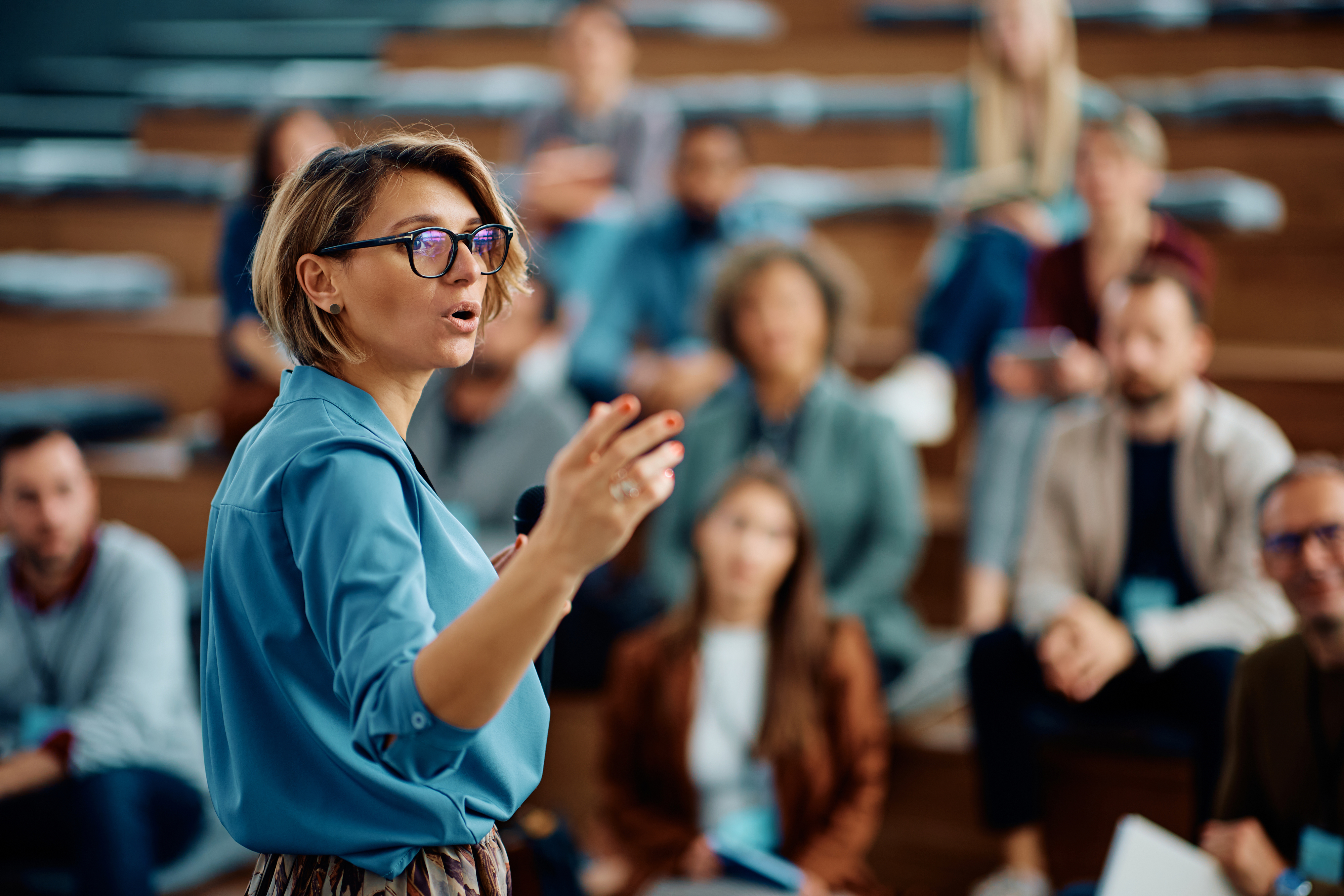 A woman in professional attire speaks passionately to a group sitting in auditorium-style seats. The image is likely from a lecture or seminar. No famous persons identified