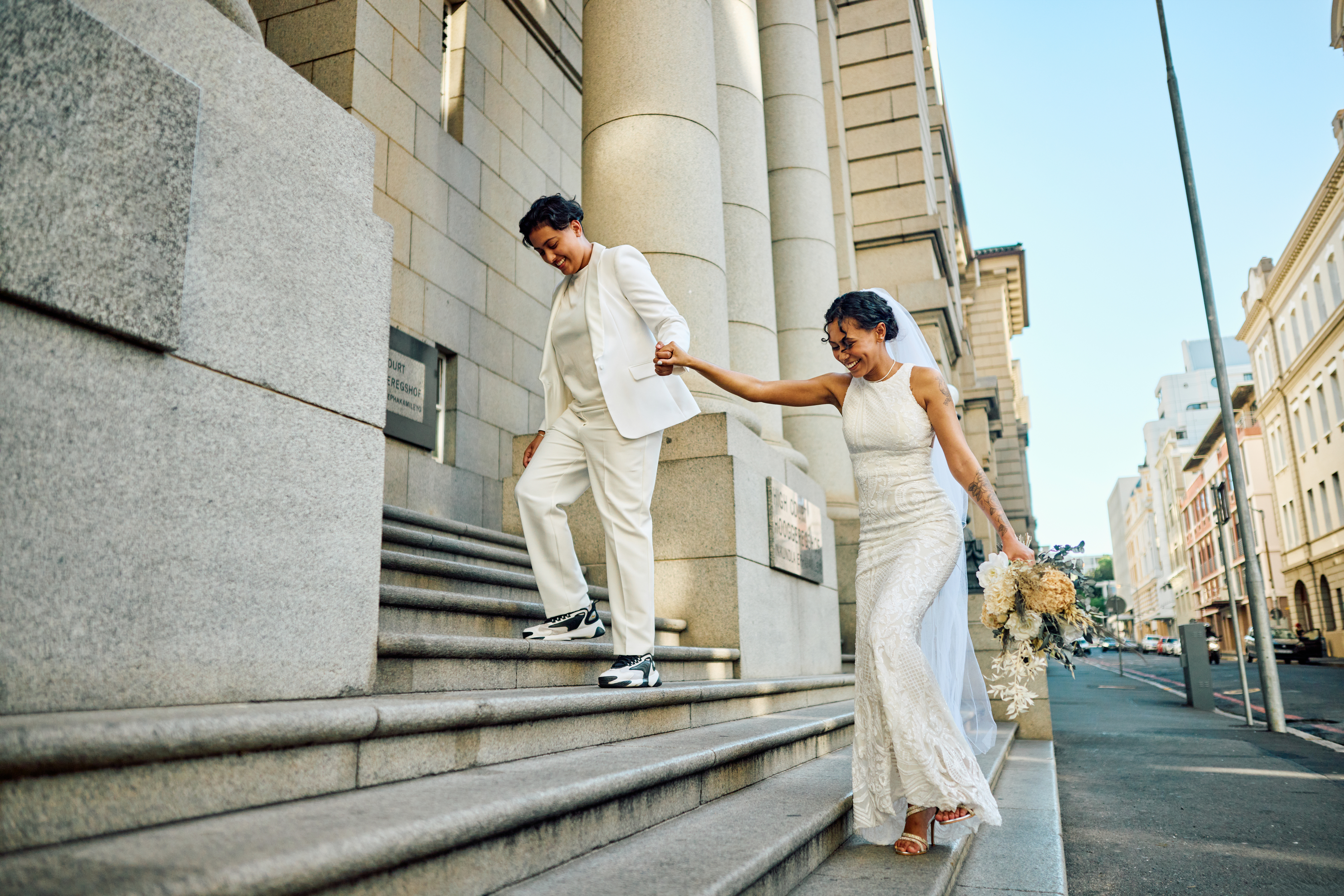 Person in a white suit and sneakers holds hands with a person in a lace wedding dress as they walk down steps outside a grand building