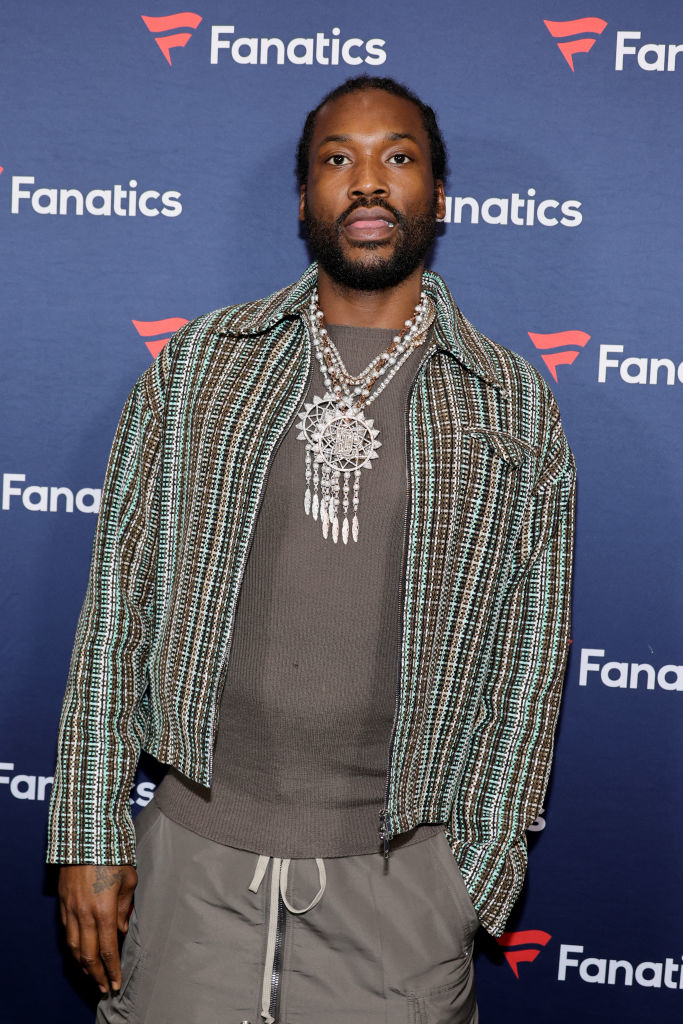 Meek Mill at a Fanatics event, wearing a beaded necklace and a checkered jacket over a gray sweatshirt