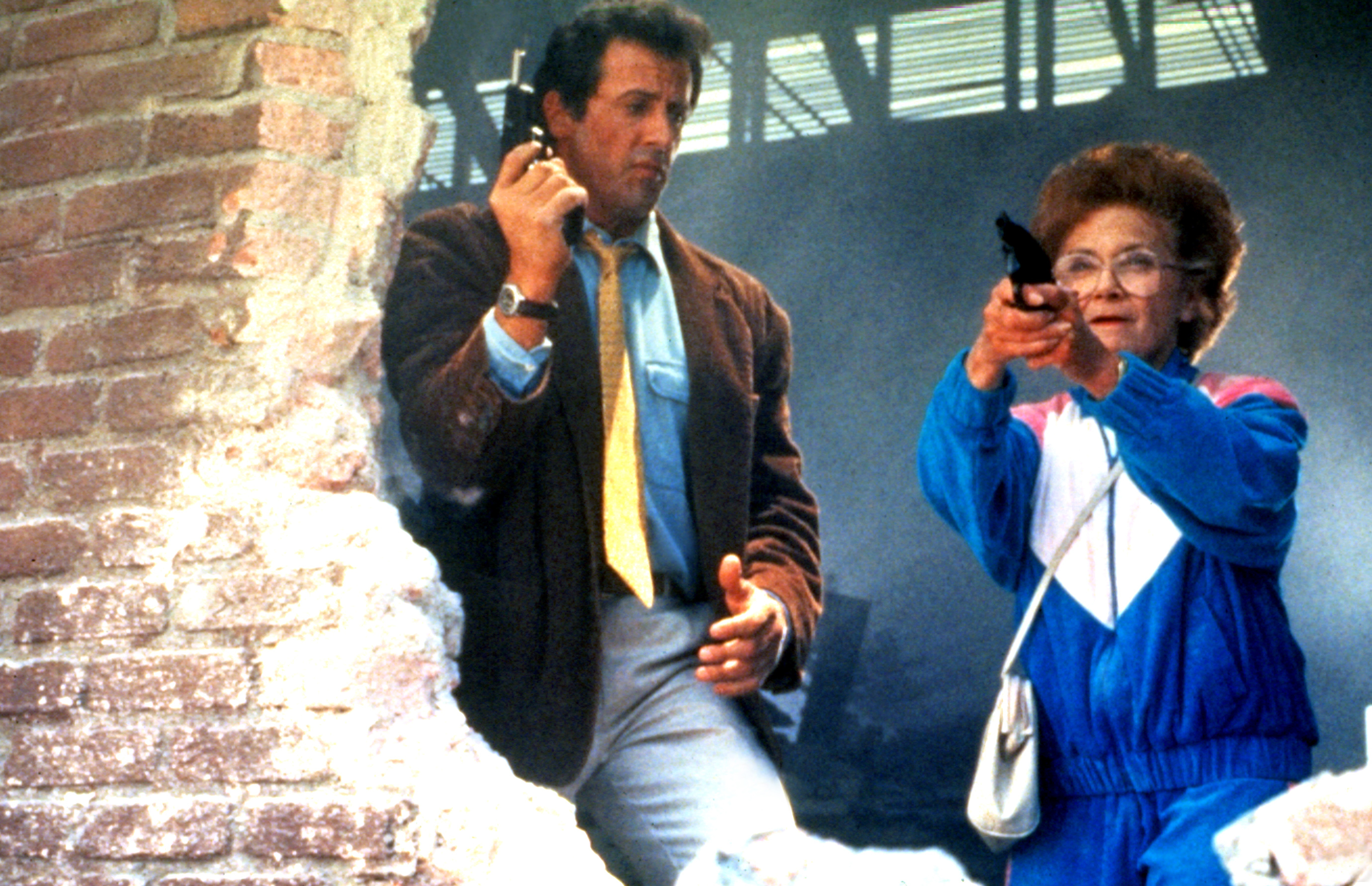 Sylvester Stallone stands next to Estelle Getty, who is aiming a gun, in a scene from "Stop! Or My Mom Will Shoot." Both are dressed in casual attire