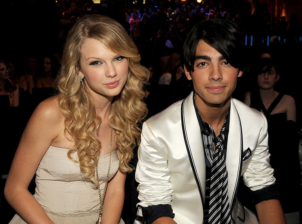 Taylor Swift in a strapless dress sits beside Joe Jonas in a white jacket with black lapels and striped tie at an event