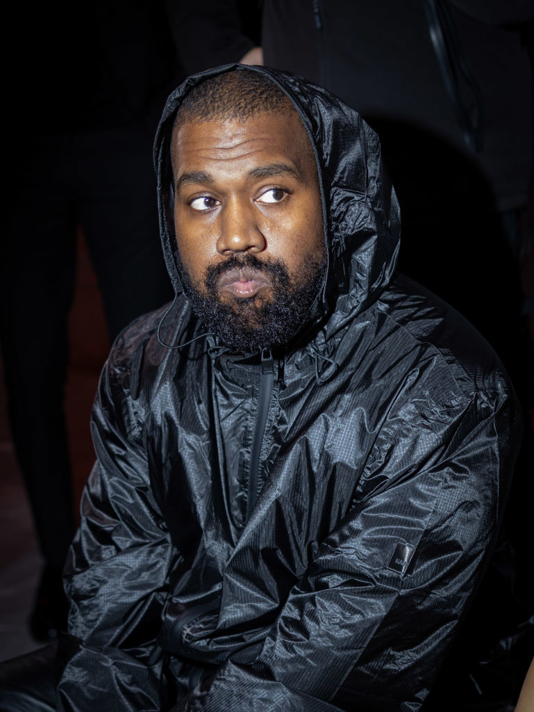 Kanye West wearing a shiny black hooded jacket, looking to the side with a neutral expression