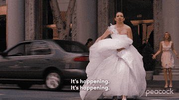 Melissa McCarthy runs in a wedding dress, holding up the skirt, while Kristen Wiig, also in a dress, stands behind her outside a building. Text reads, &quot;It's happening.&quot;