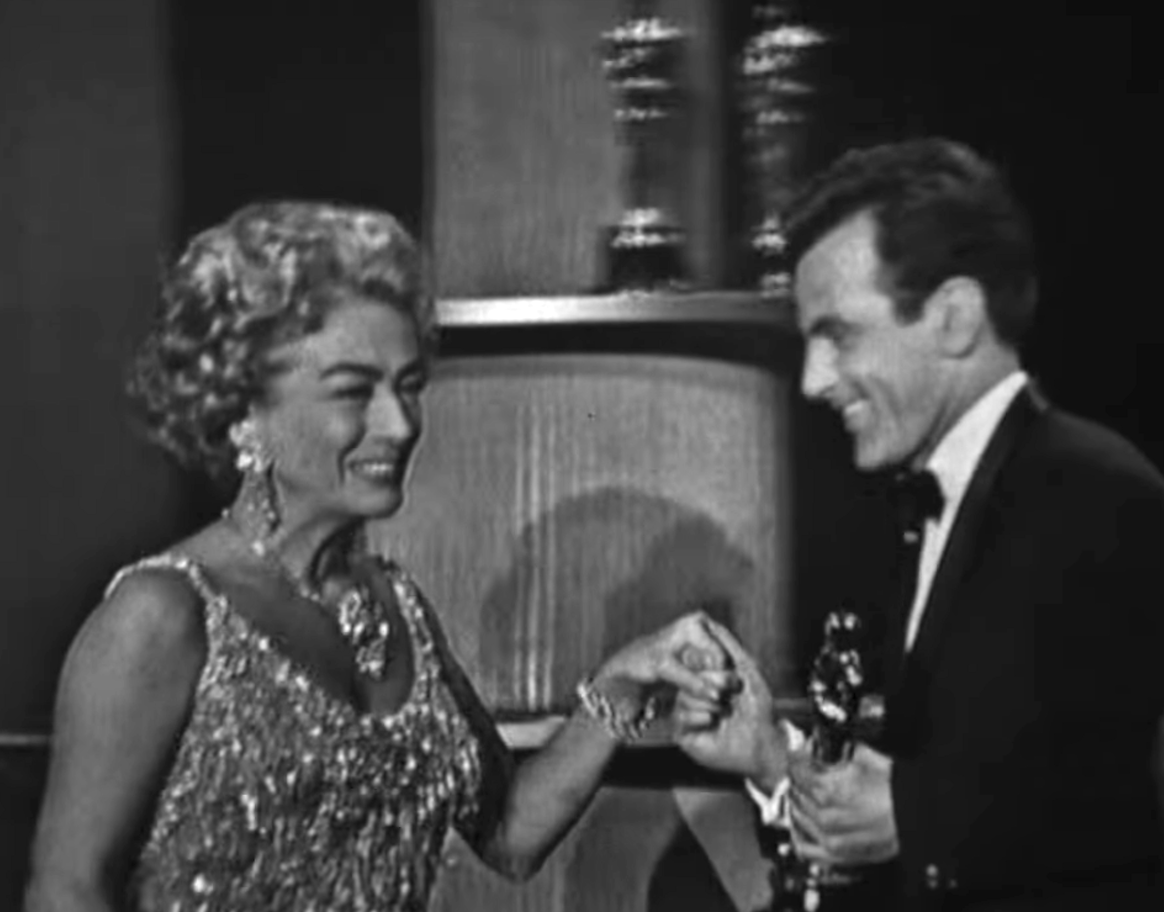 Joan Crawford, in an embellished gown and ornate jewelry, holds hands with a man in a tuxedo holding an Oscar trophy on stage