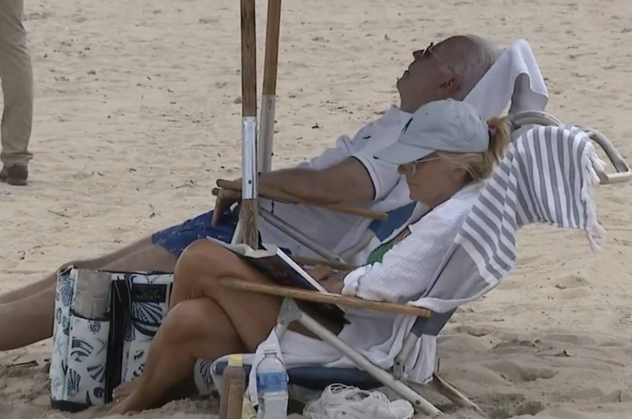 Joe Biden and Jill Biden sit on lounging chairs under an umbrella on the beach, Joe resting and Jill reading a book