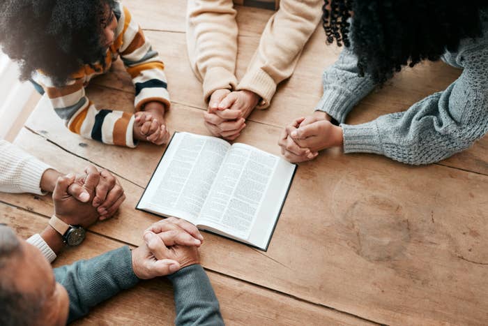 Six individuals hold hands around an open Bible on a wooden table