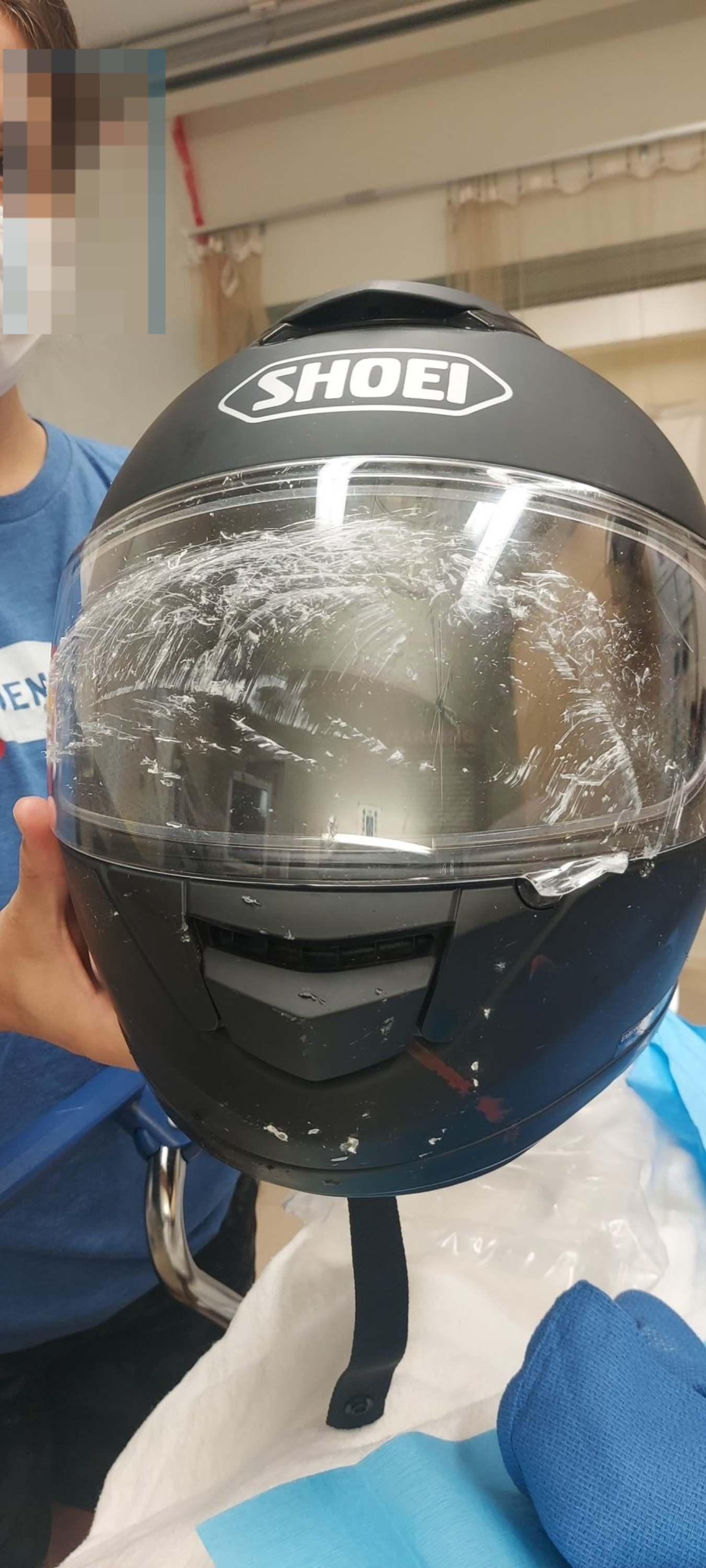 A person holds a worn motorcycle helmet with visible scratches on its visor. The helmet has the brand name "Shoei" on the front