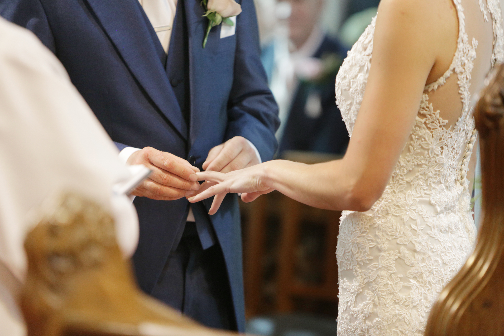 A groom places a ring on the bride's finger during their wedding ceremony. Both are dressed in formal wedding attire. Only their torsos and hands are visible