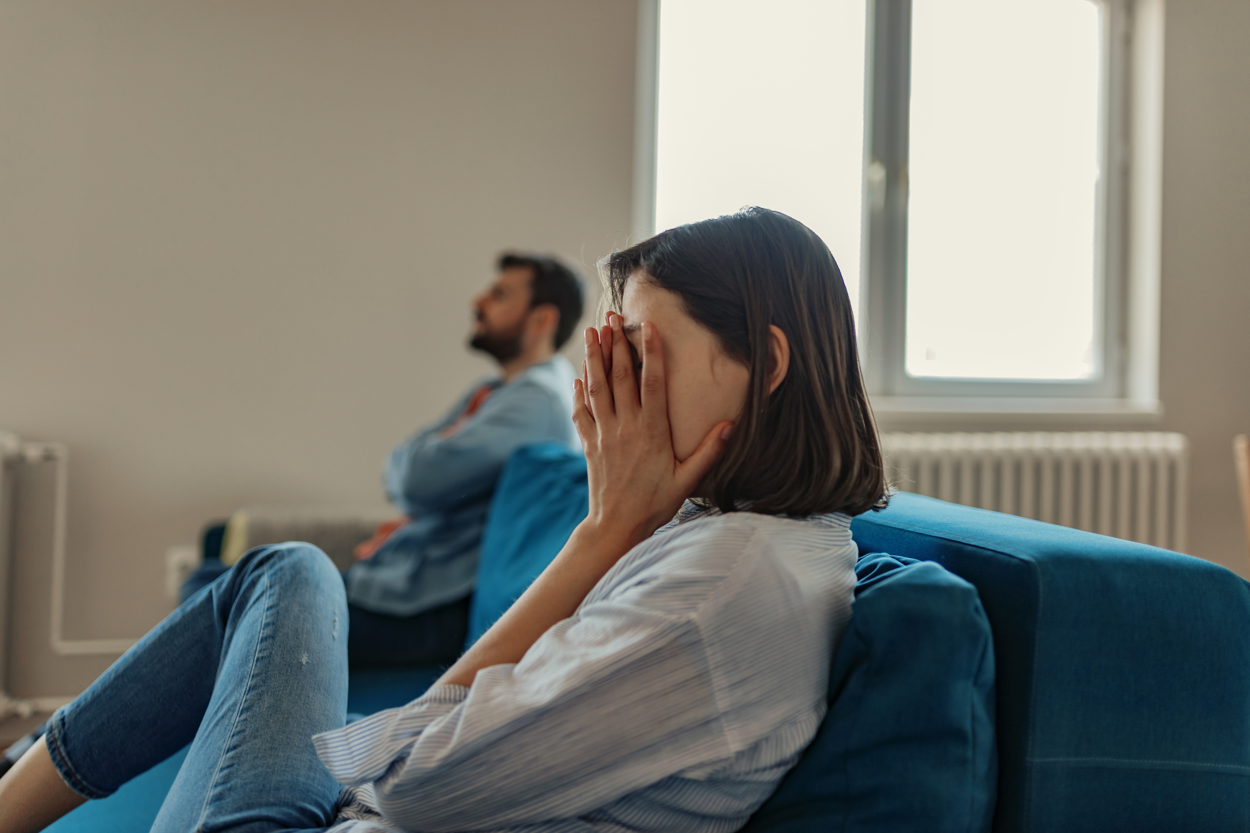 A woman sits on a couch with her face in her hands, looking distressed. A man sits in the background with his arms crossed, looking away