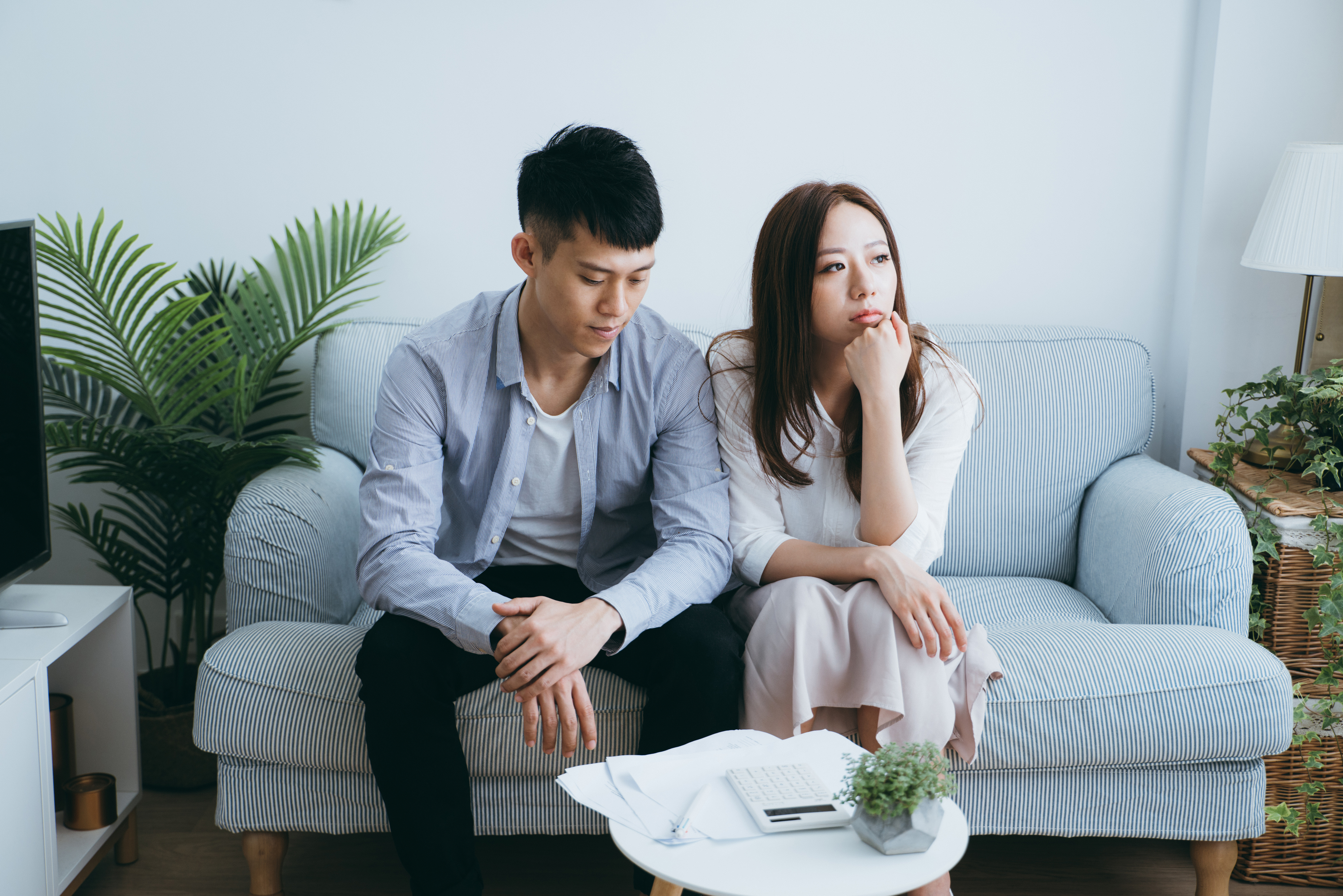 A couple, a man and woman, sit on a couch looking contemplative. The woman rests her chin on her hand while the man looks down