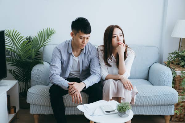 A couple, a man and woman, sit on a couch looking contemplative. The woman rests her chin on her hand while the man looks down