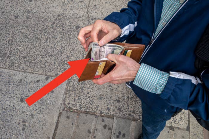 Person in a checkered shirt and jacket taking cash out of a tan wallet while standing on a sidewalk