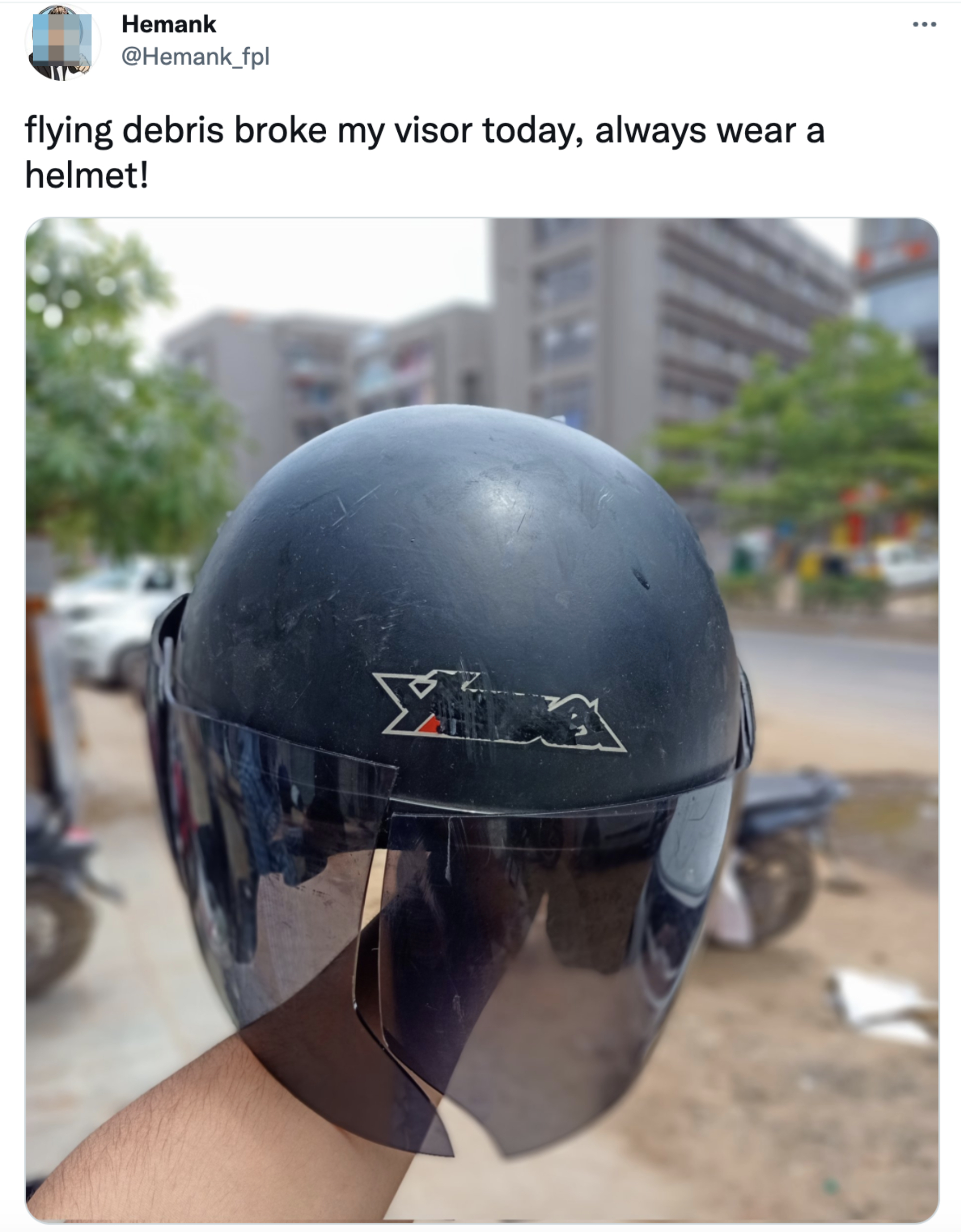 A hand holds a black helmet with a transparent visor in an outdoor setting. Text above reads: "flying debris broke my visor today, always wear a helmet!"