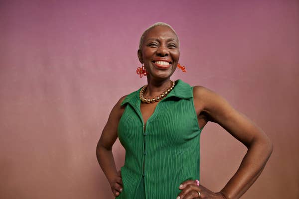 A smiling woman poses confidently against a simple background. She is wearing a sleeveless green dress and bold red earrings