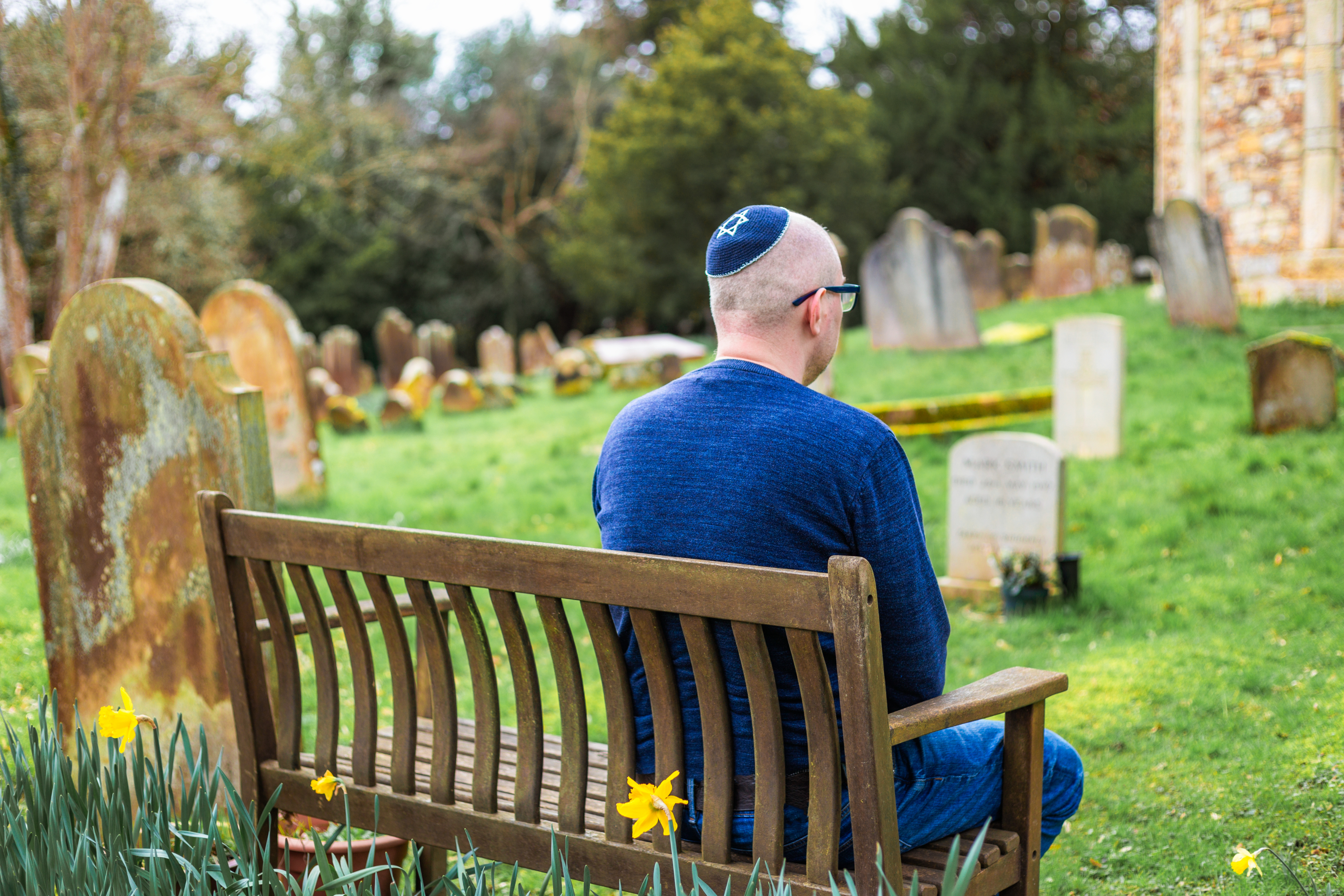 A man wearing a kippah sits on a bench in a cemetery, facing away, looking at gravestones. The cemetery has many old tombstones and blooming daffodils