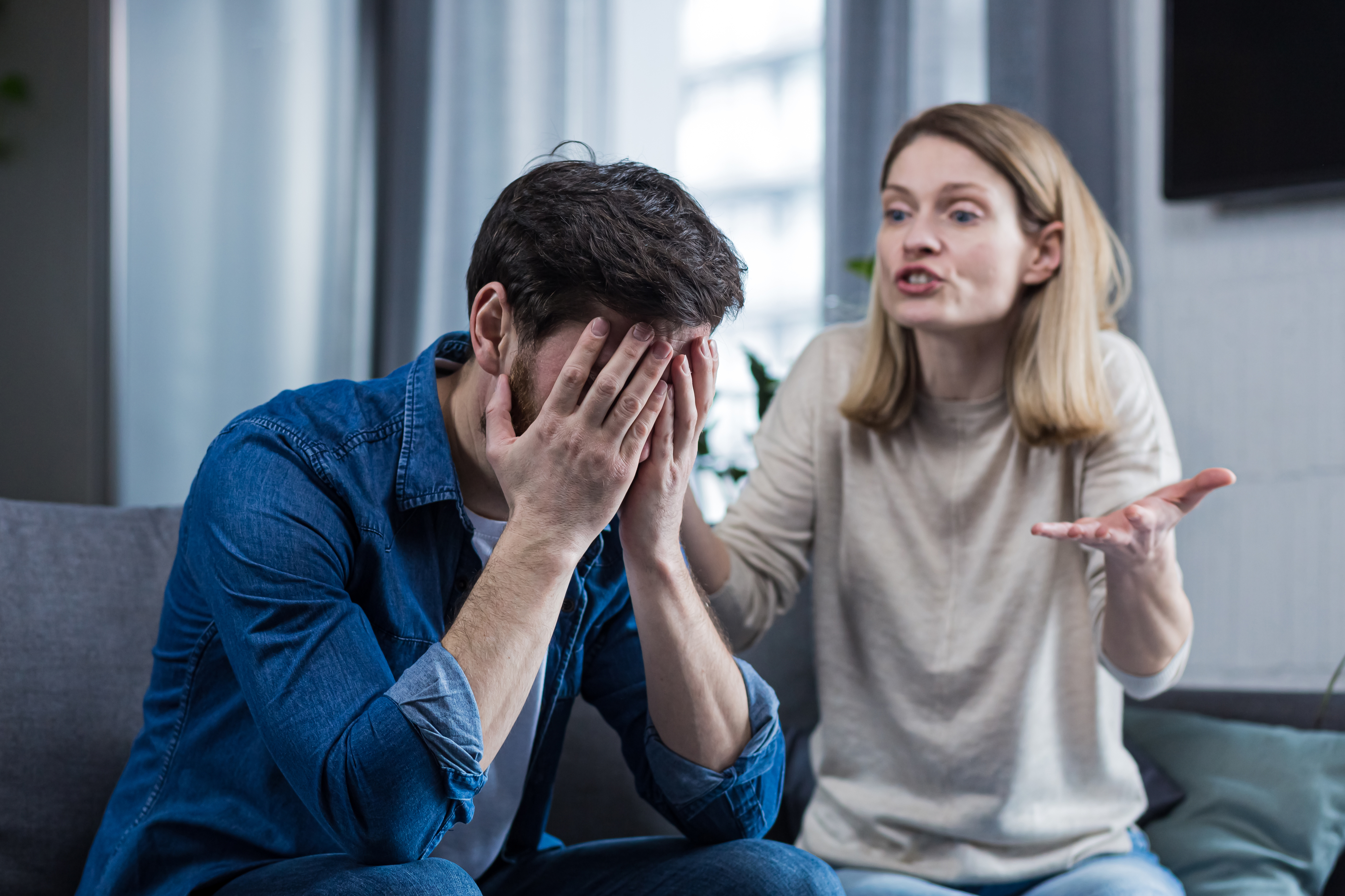 A man sits with his hands covering his face, appearing distressed, while a woman beside him gestures, seemingly arguing or expressing frustration