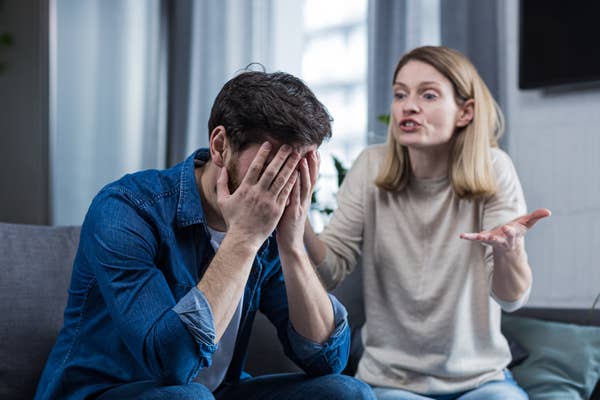 A man sits with his hands covering his face, appearing distressed, while a woman beside him gestures, seemingly arguing or expressing frustration