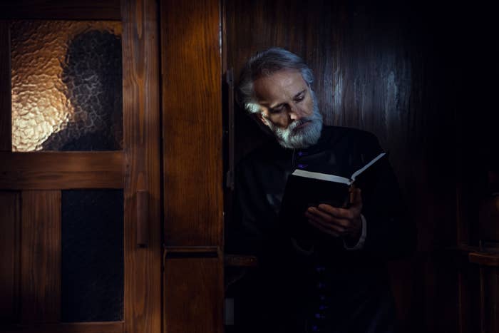 An elderly man with gray hair and beard reads a book while standing in a wooden confessional booth, appearing contemplative