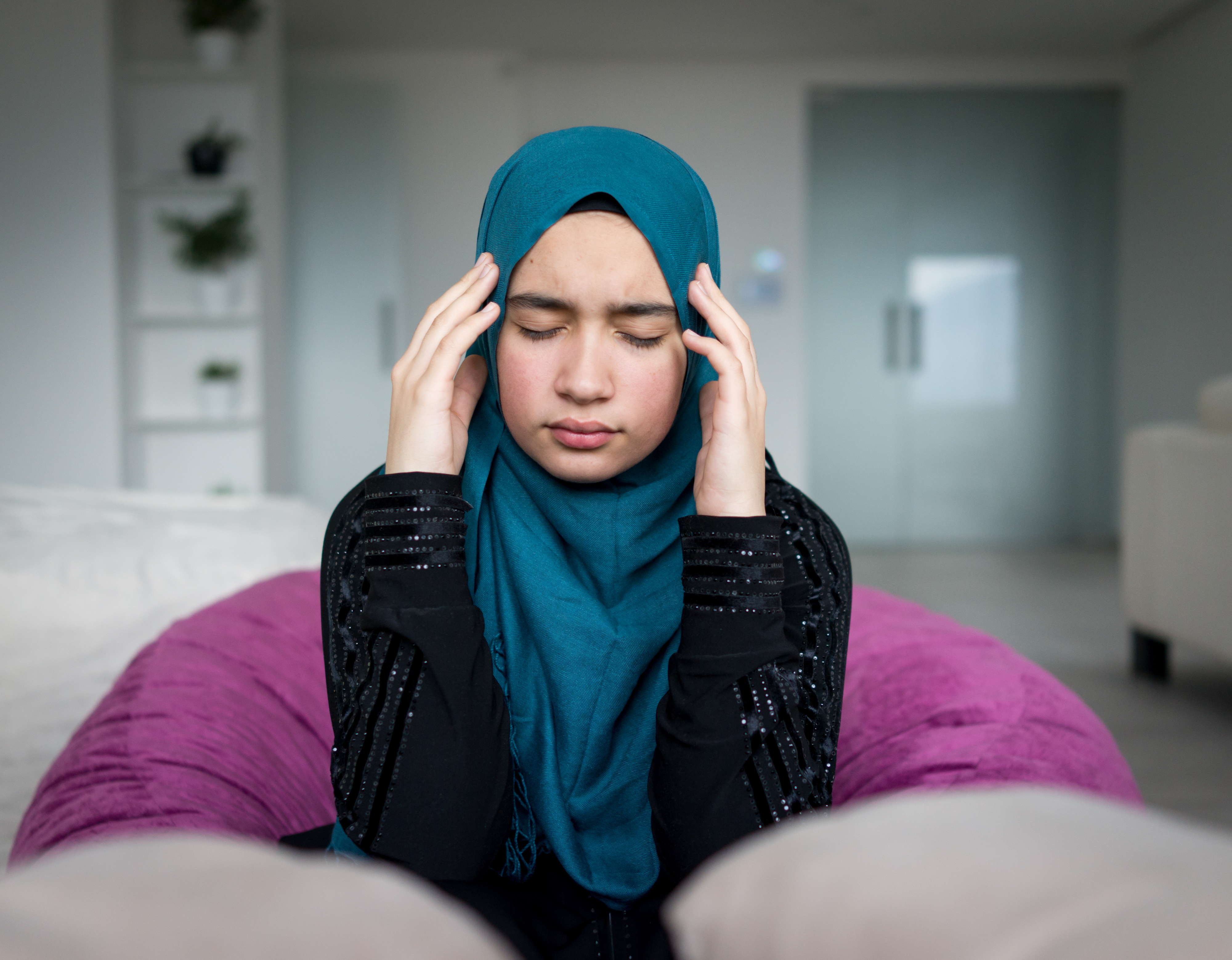 A woman wearing a hijab sits on a couch with her eyes closed and hands on her temples, looking distressed