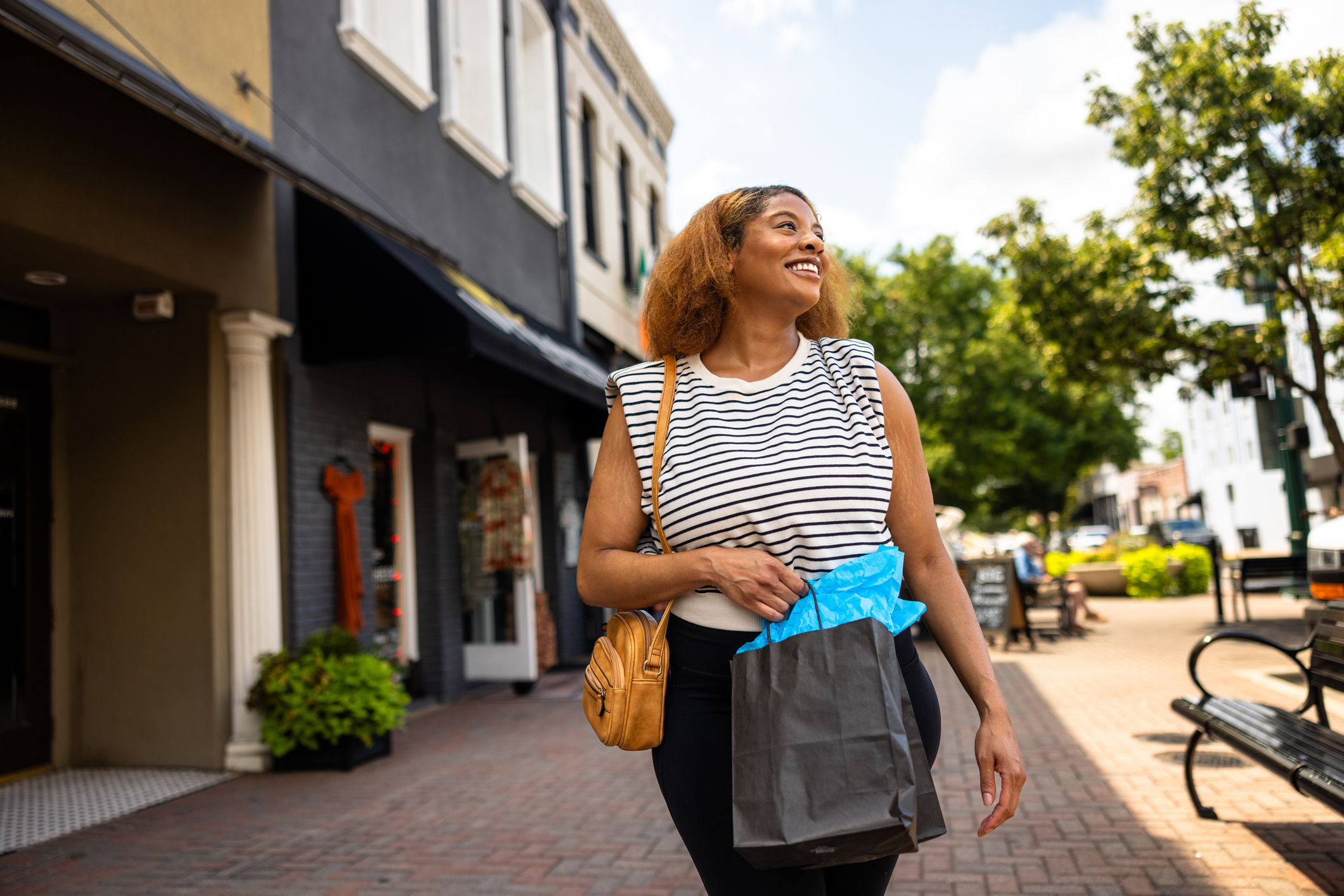 Person walking through a shopping district, smiling, and holding a shopping bag with crumpled blue paper visible