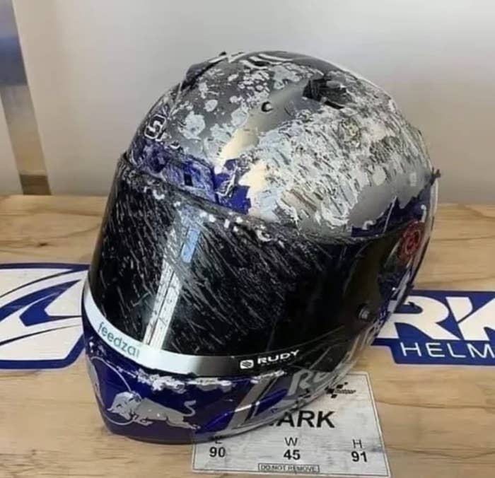 A racing helmet with a scratched visor and damage across the top is displayed on a table. The helmet has branding from Shark, Red Bull, Rudy, and Feebzal