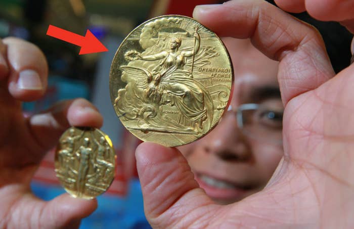 A person holds up two Olympic gold medals, showing the intricate designs on the front. Their face is partially visible behind the medals