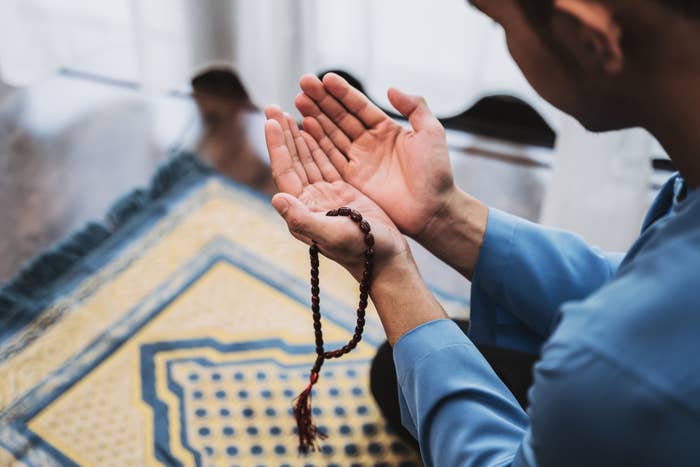 A person in prayer, holding prayer beads, with a prayer rug in front of them. The person is wearing a light-colored shirt and has their hands raised
