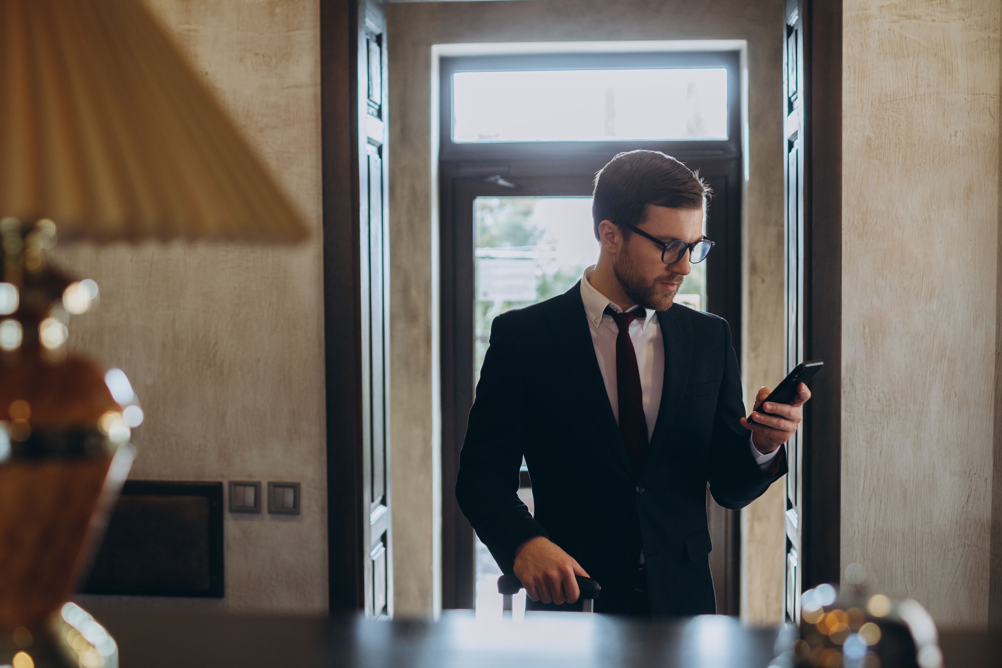 A man in a business suit with glasses is looking down at his phone while holding a suitcase in a modern lobby or reception area