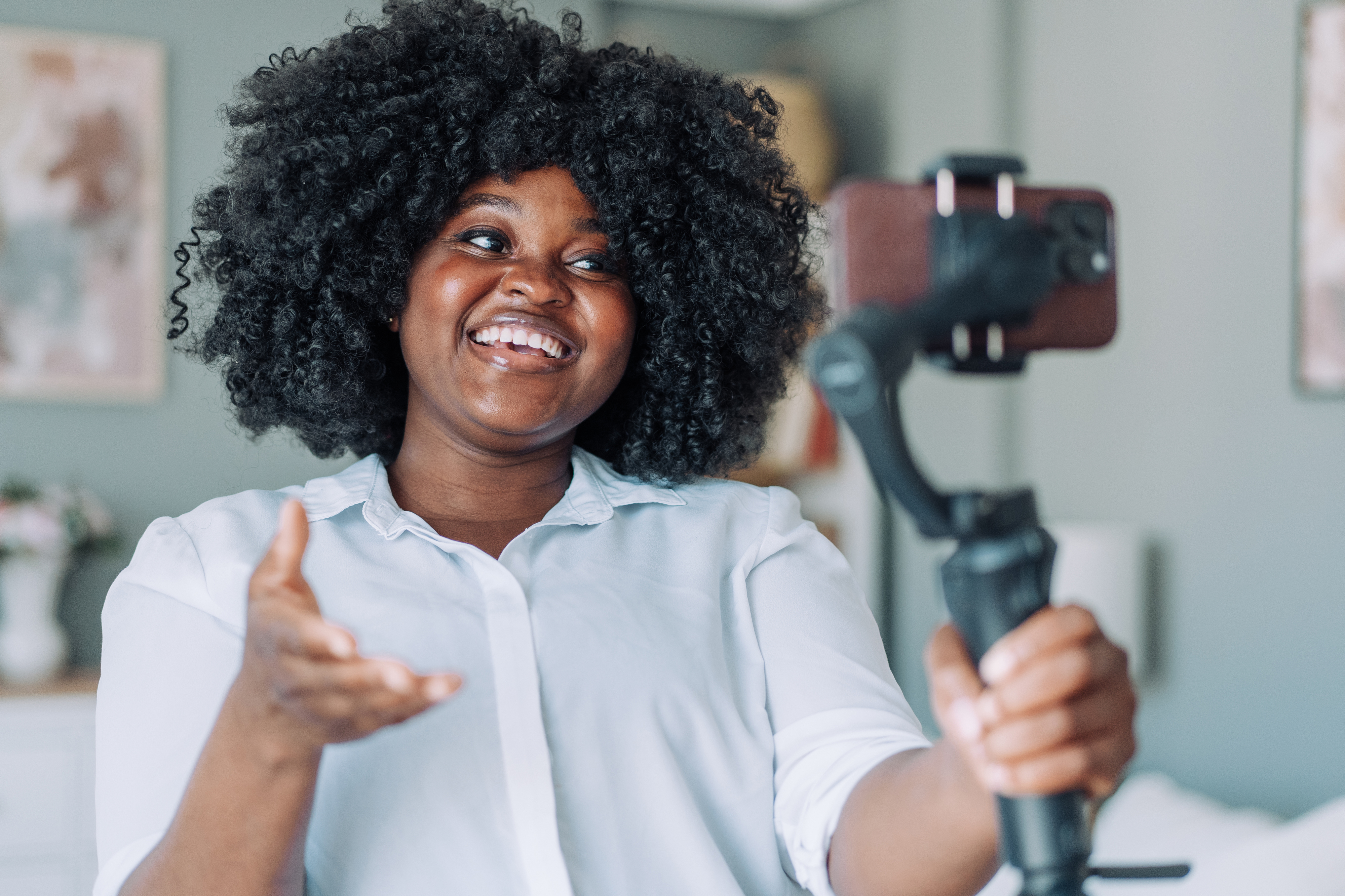Woman with curly hair smiles and gestures while recording herself with a smartphone on a tripod indoors